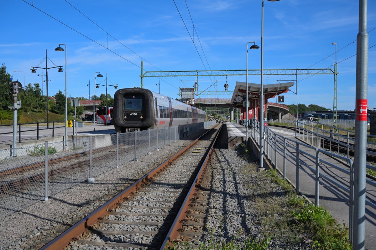 En bild tagen vid en tågstation med ett tåg som står stilla på en av spåren. I bakgrunden syns en buss, samt övergångar för bilar och en del grönska. Stationen har en röd byggnad och en ramp med räcken. En solig dag med klarblå himmel.