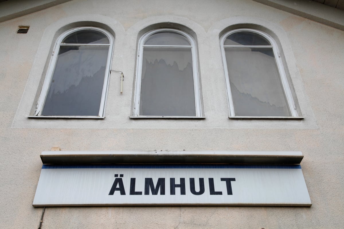 En fasad på en ljus byggnad med tre välvda fönster på övervåningen. Under fönstren sitter en skylt med texten "ÄLMHULT" i stora svarta bokstäver. Fasaden är putsad och verkar vara i behov av underhåll.