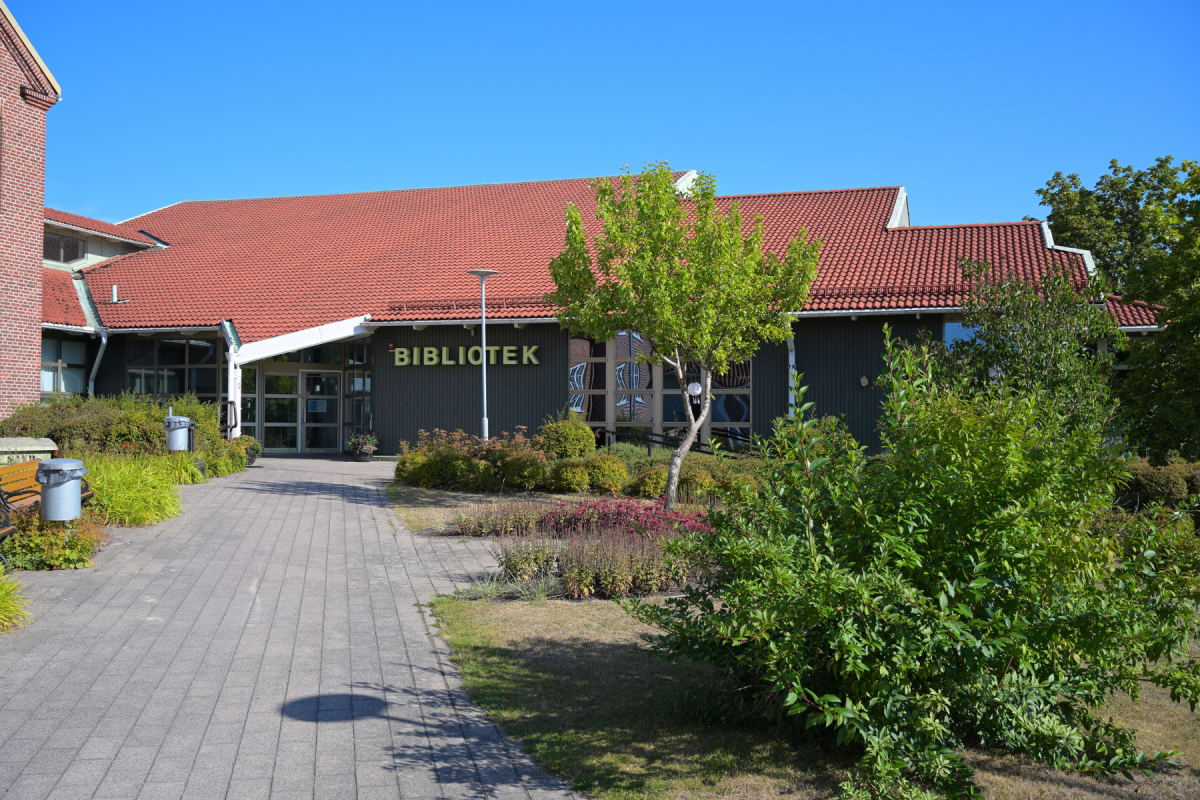 En fasadvy av en enplansbyggnad med rött tegeltak och mörk fasad med texten "BIBLIOTEK" i ljusa bokstäver. Framför byggnaden finns en gångväg i stenplattor, gräsmattor och buskar. Himlen är klarblå.