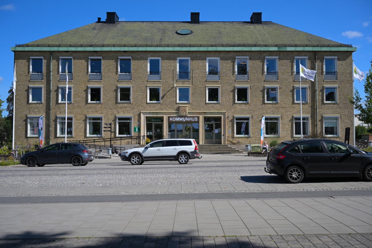 En bild visar ett kommunhus med tegelväggar och ett grönt tak under en blå himmel. Framför byggnaden syns tre bilar parkerade, samt en person som sitter på en bänk. Två flaggor med texten "Älmhults kommun" syns.