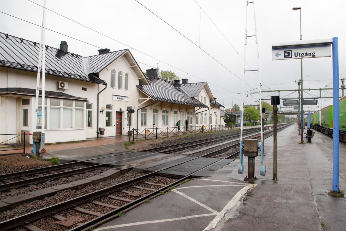 En bild som visar en järnvägsstation med spår, perronger och vita stationsbyggnader. På en av byggnaderna står "ÄLMHULT" och en skylt med "Utgång" och en pil är synlig. Ett godståg står vid en perrong och några människor syns på perrongen.