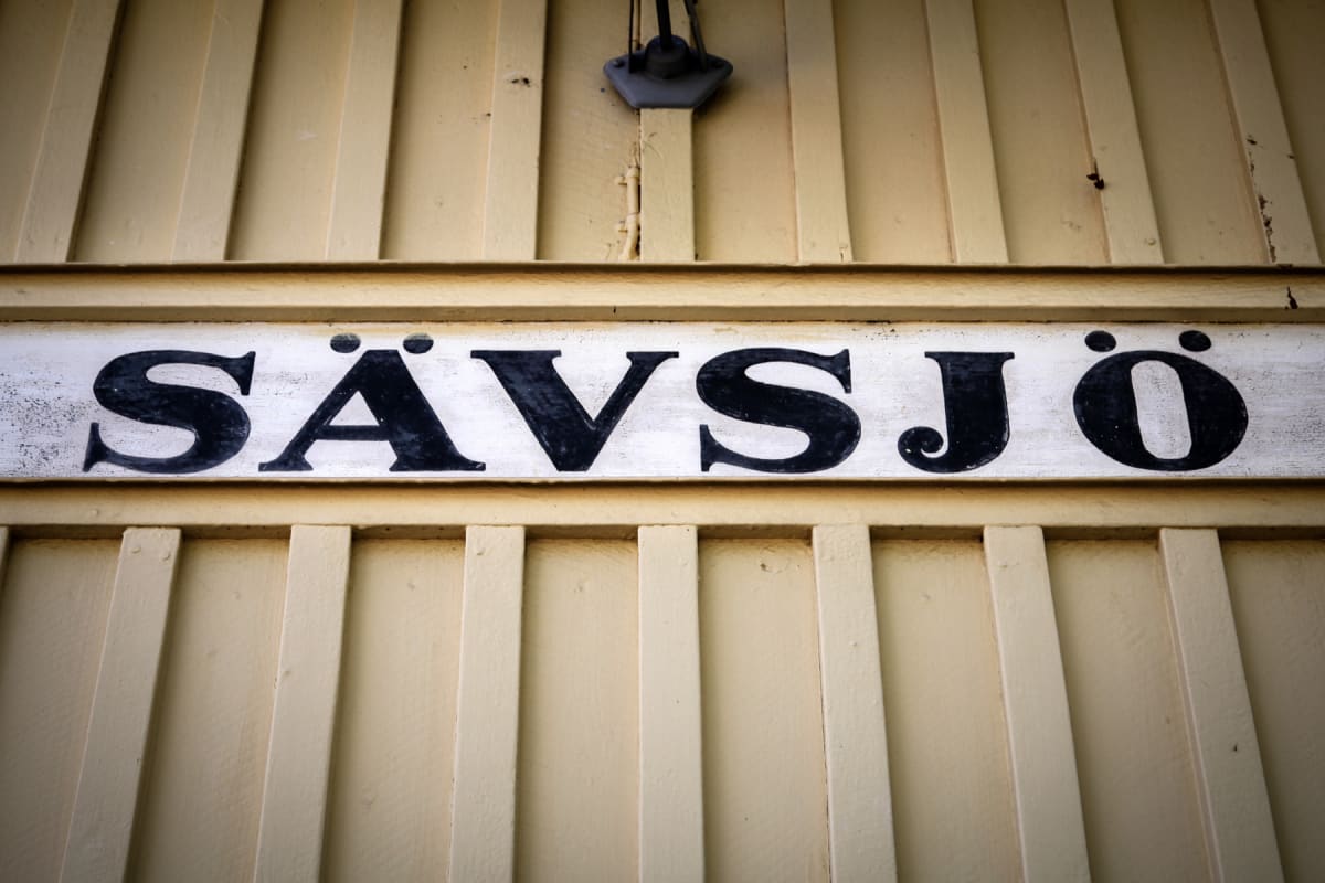 En gulmålad träbyggnad med en vit skylt med svarta bokstäver som stavar "SÄVSJÖ" i mitten. Ovanför skylten sitter en mörk lampa eller fäste på träväggen.