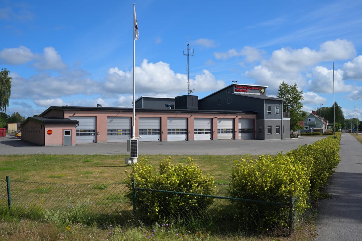 En enplansbyggnad med flera garageportar och en tillbyggd del med två våningar under en blå himmel med vita moln. Byggnaden har rosa väggar med mörkgråa detaljer och ett skyltfönster med texten "Räddningstjänsten Hultsfred".