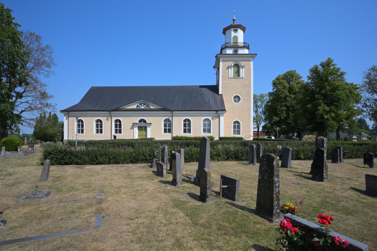 En gul kyrka med ett svart tak och ett torn med en grön kupol och klocka, omgiven av en kyrkogård med grå gravstenar och vissnat gräs. En grön häck löper längs kyrkans framsida och träd syns i bakgrunden. En klarblå himmel utgör bakgrunden.