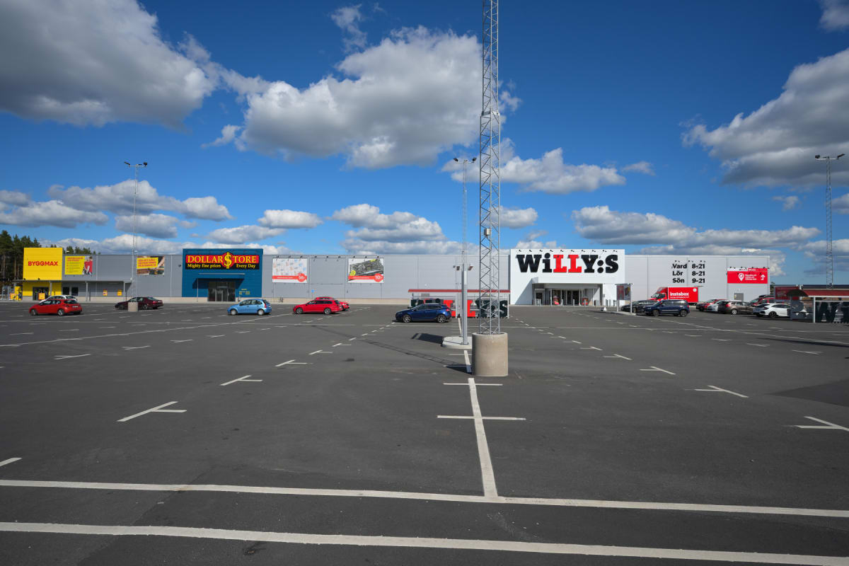 En stor, asfalterad parkeringsplats med målade körbanor och markerade parkeringsrutor. I bakgrunden syns en lång byggnad med flera butiker, inklusive "BYGGMAX", "DOLLARSTORE" och "WILLY:S". Flera bilar är parkerade på platserna och några kör på parke