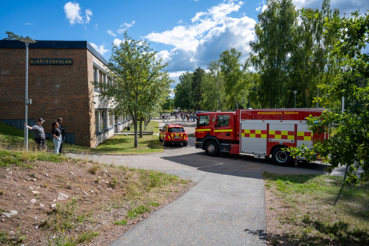 Bilden visar ett räddningsfordon och en röd personbil parkerade på en asfalterad yta framför en tegelbyggnad märkt "ALBÄCKSSKOLAN". I bakgrunden syns flera människor samlade, samt träd och en klarblå himmel med moln. Tre personer står närmare byggnad
