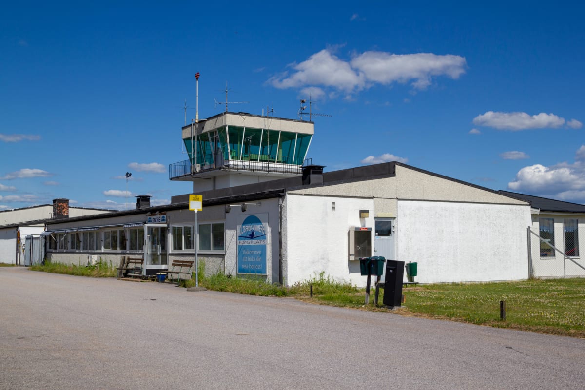 En vit byggnad med ett grönrutigt flygledartorn under en blå himmel med vita moln. Byggnaden har fönster, en "Entré"-skylt, ett par bänkar utanför samt en skylt med texten "FLYGPLATS". Två brevlådor och en telefonkiosk syns också.
