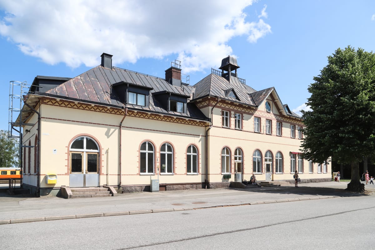 En stor beige byggnad med tak i olika nivåer ses under en sommarhimmel med vita moln och blå himmel. Framför byggnaden finns en asfalterad yta med en gata närmast betraktaren och grus längre bort, samt ett träd med gröna löv.