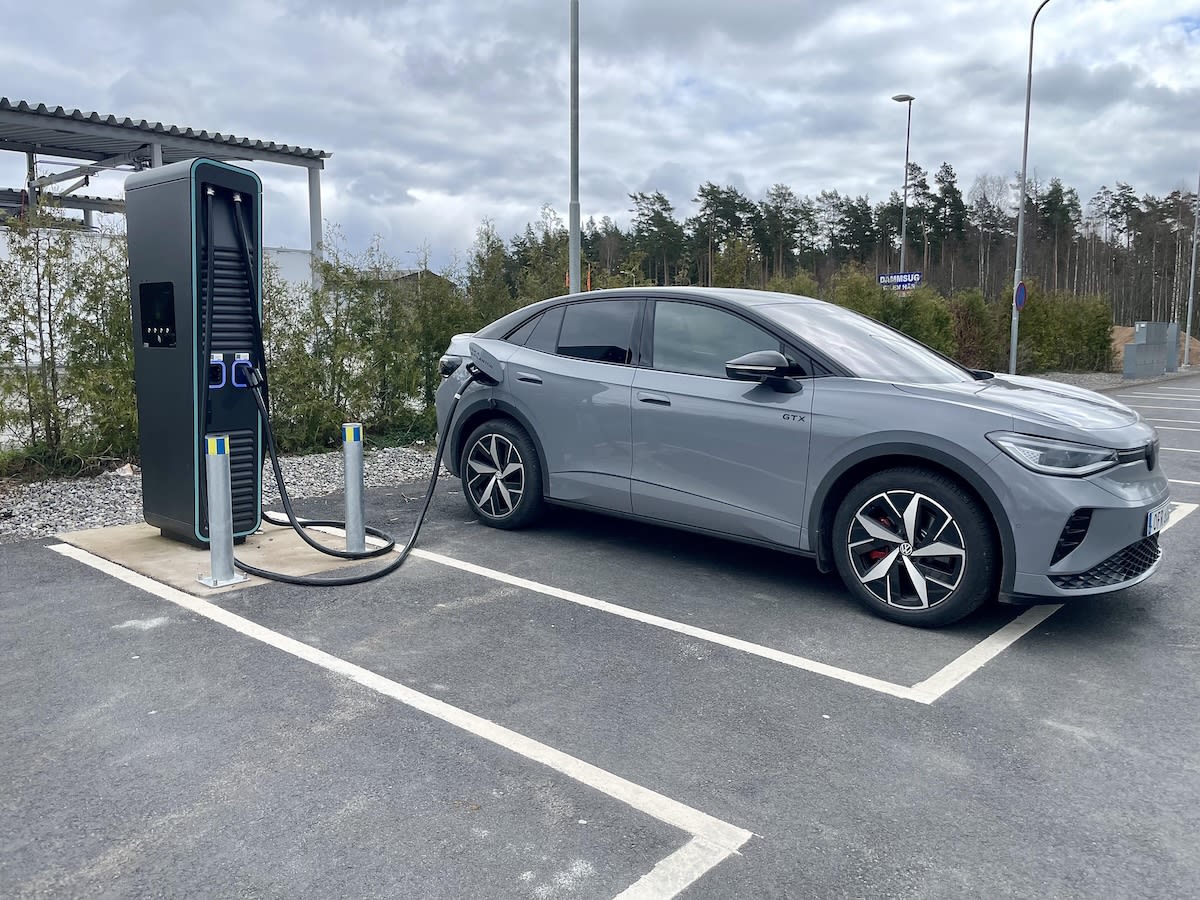 En grå elbil av typen Volkswagen ID.4 GTX står parkerad vid en laddstation utomhus under en gråmulen dag. Laddkabeln är ansluten till bilens vänstra sida. I bakgrunden syns skog och en skylt med texten "DAMMSUG".