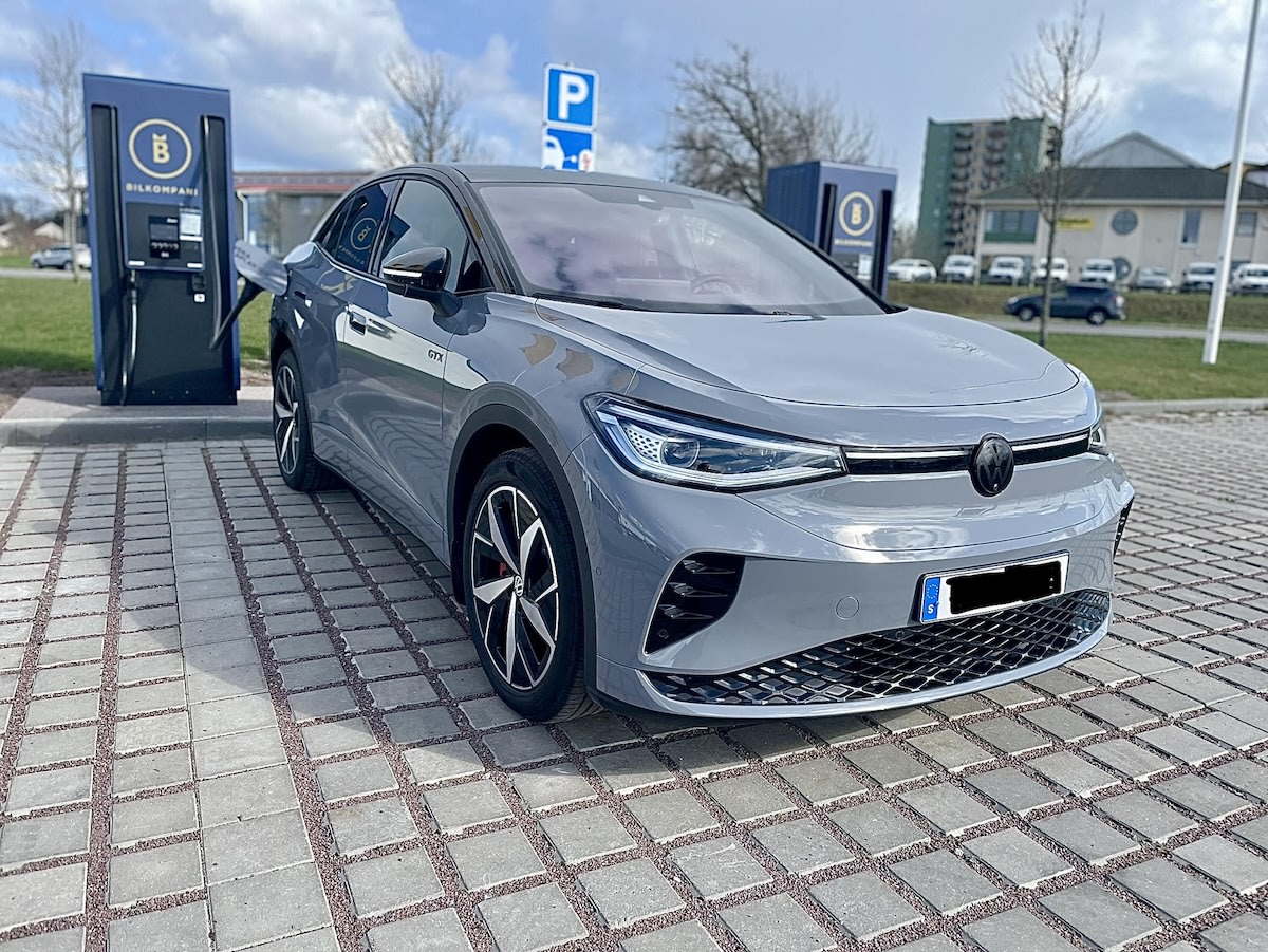 En grå bil av märket Volkswagen laddas vid en laddstation utomhus. Bilen står parkerad på en yta av stenplattor med grus mellan. I bakgrunden syns andra byggnader och träd med kala grenar.