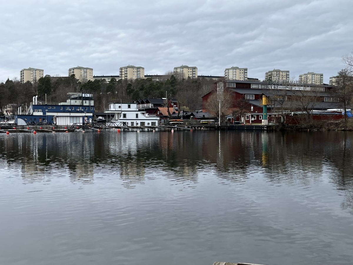 En bild som visar en småbåtshamn och dess omgivning. I förgrunden syns vatten med bryggor och bojar. Längs landkanten står olika byggnader, inklusive en blå och vit husbåt, röda byggnader och en skylt där det står "PAMPAS MARINA".