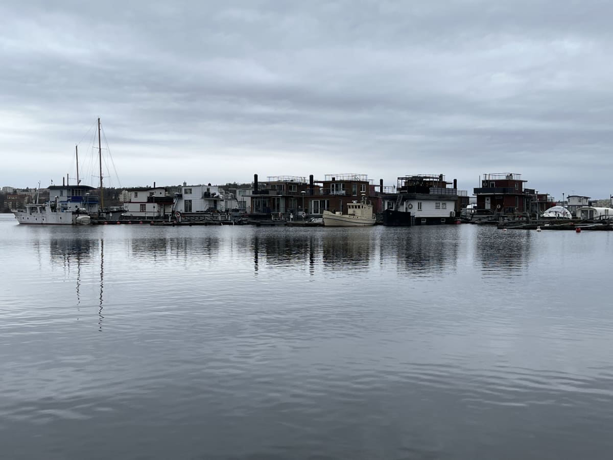 En bild som visar en rad med husbåtar och båtar som ligger förtöjda längs en lugn vattenyta under en molnig himmel. Byggnaderna har olika utseenden, några med vita och andra med mörka fasader. I förgrunden reflekteras byggnaderna och masterna från en