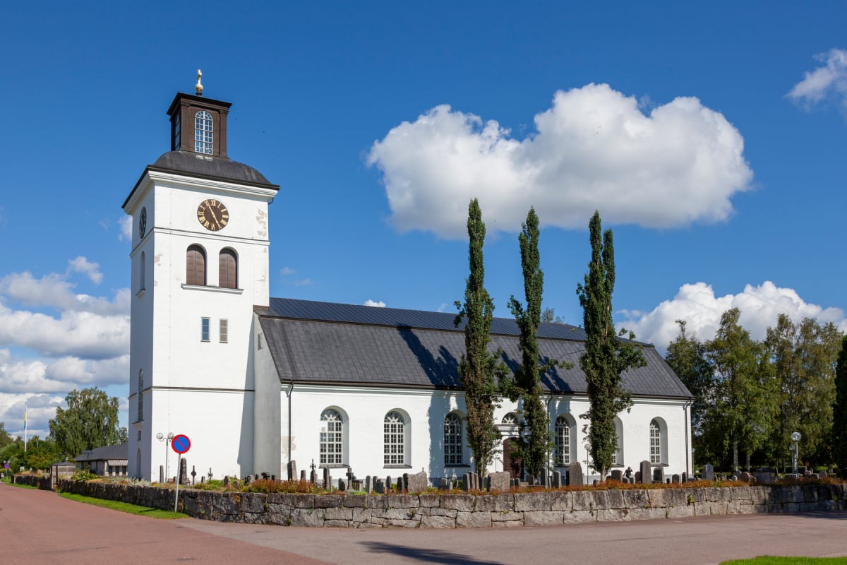 En vit kyrka med klocktorn och svart tak under en klarblå himmel med vita moln. Framför kyrkan finns en grå stenmur, en kyrkogård med gravstenar, höga träd och en asfalterad väg. Ett "parkering förbjuden"-märke syns även. Kyrkan har flera fönster och