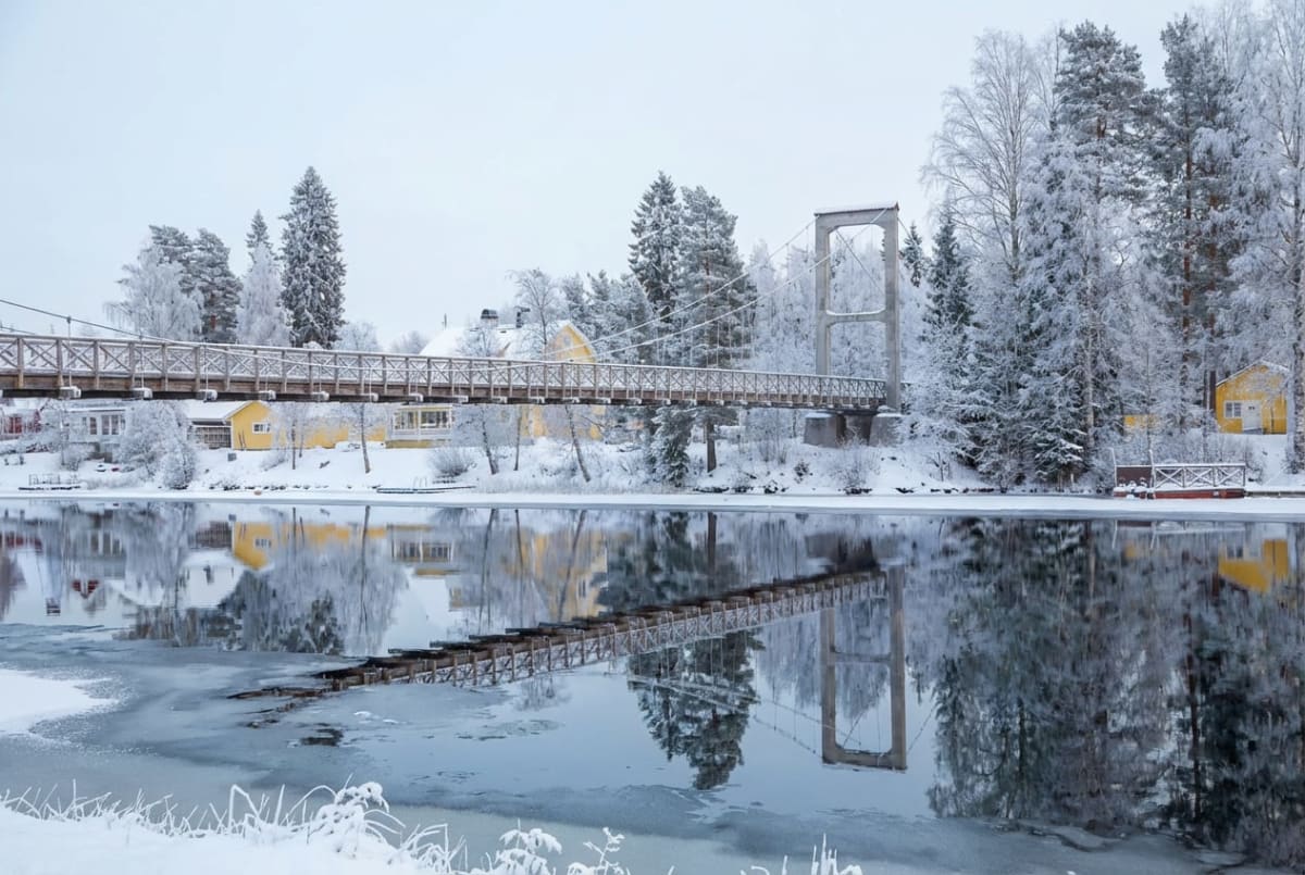 En vinterbild som visar en hängbro en bit ovanför ett delvis isbelagt vattendrag. På vardera sida av vattendraget står snötäckta träd och mark. Flera gula hus syns på den bortre stranden. Bron och omgivningen reflekteras tydligt i vattnet.