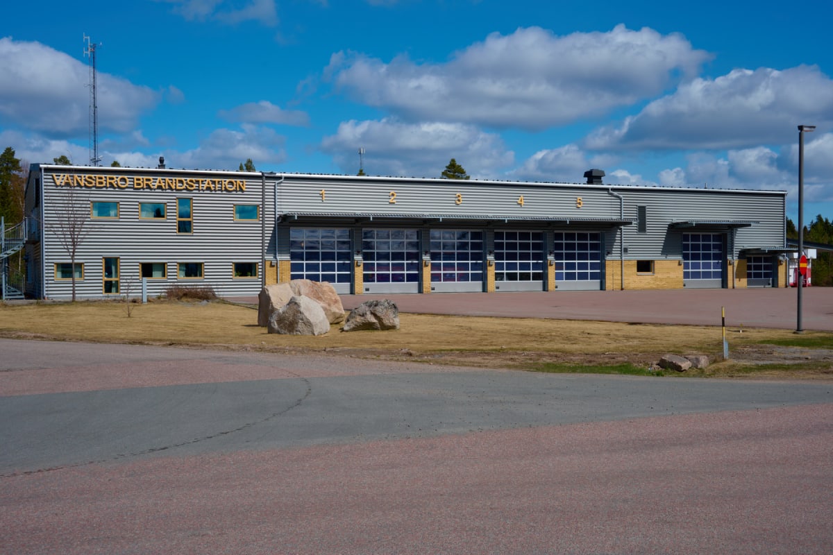 En modern brandstation med grå korrugerad plåtfasad och ett stort antal portar med fönster. Entrén har texten "VANSBRO BRANDSTATION" i guldiga bokstäver. Framför byggnaden finns en asfalterad yta, en gräsmatta och ett par stora stenar. En klarblå him