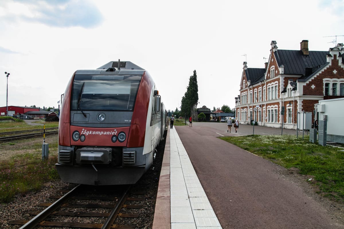 En röd och vit tågvagn står vid en perrong bredvid en stor tegelbyggnad. I bakgrunden syns skog och en molnig himmel. Flera personer syns på perrongen och framför byggnaden. På byggnaden skyltas namnet ''Tågkompaniet'', en text som även syns på tåget
