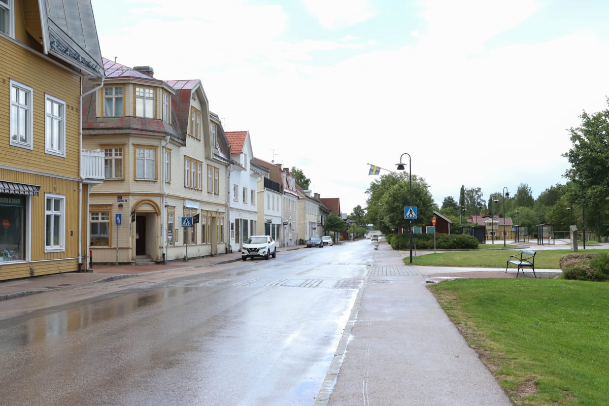 En gatsvy över en stadsgata med byggnader längs ena sidan och en gräsyta med busshållplatser på den andra. Gatan är våt, vilket reflekterar himlen. Flera bilar är parkerade längs gatan, och trafikskyltar syns intill vägen.