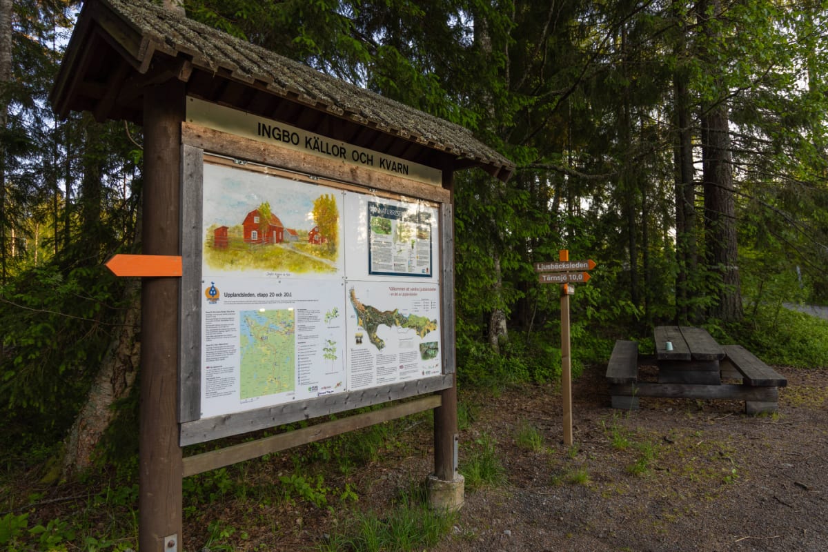 En informationstavla vid en vandringsled i en skogsmiljö. På tavlan syns en illustration av röda byggnader, kartor och text. Bredvid tavlan står en skylt för 'Ljusbäcksleden' och en bänk med bord. Marken består av grus och gräs och i bakgrunden syns