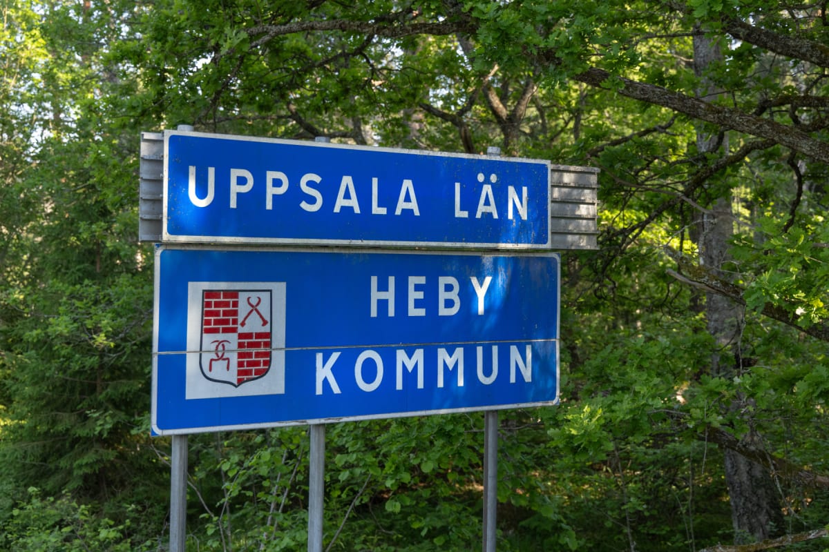 En blå vägskylt med texten "UPPSALA LÄN" överst, och "HEBY KOMMUN" under, samt ett kommunvapen bredvid texten. Skylten står utomhus omgiven av grönskande träd, buskar och lövverk på en solig dag.