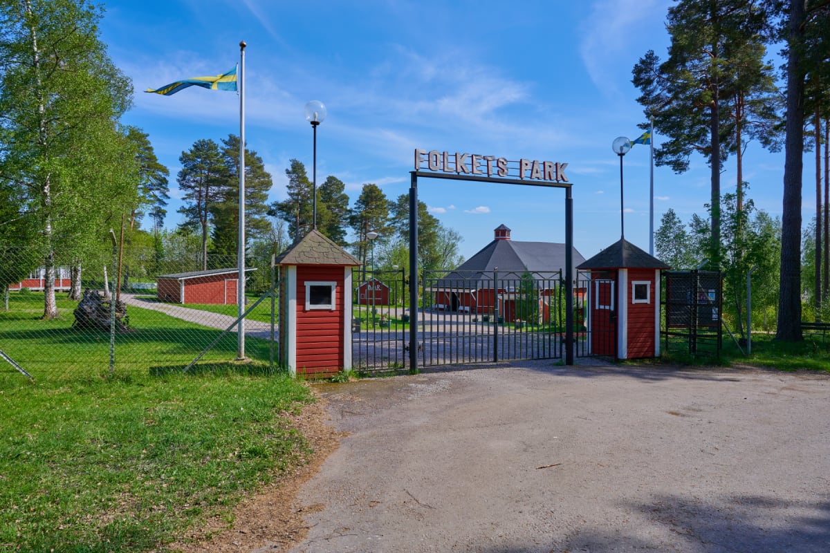 En väg leder fram till en järngrind med texten "FOLKETS PARK" ovanför. Två röda kurer står på var sin sida om grinden. Bakom grinden syns flera röda byggnader, träd och en gräsmatta. Två svenska flaggor vajar på flaggstänger.