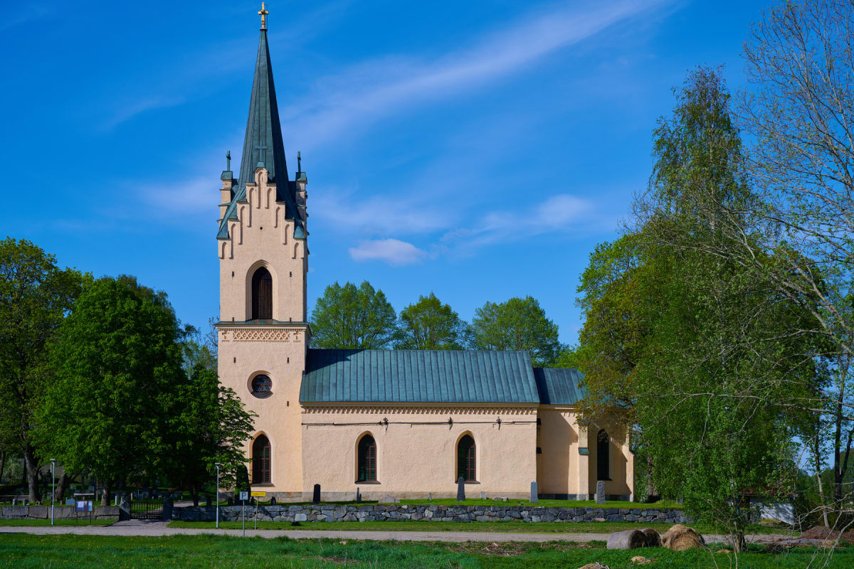 En gul kyrka med en hög, spetsig spira står under en klarblå himmel. Runt kyrkan syns grönskande träd och en gräsmatta. En stenmur avgränsar kyrkogården.