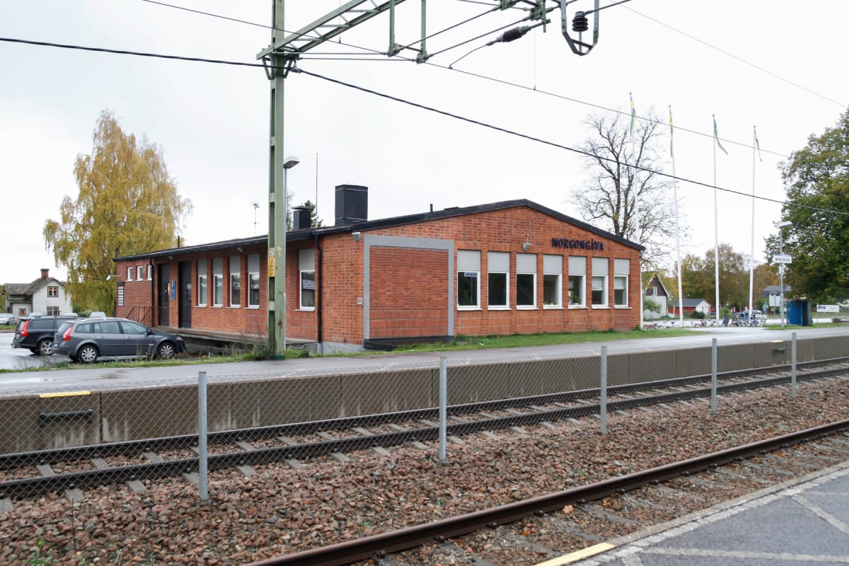 En tegelbyggnad vid järnvägsspår under en molnig höstdag. Två bilar, varav en mörk kombibil, står parkerade på en asfalterad yta nära byggnaden. En stor ljusgul björk till vänster och träd med grönområden runtom syns. Skylten på byggnaden säger "MORG