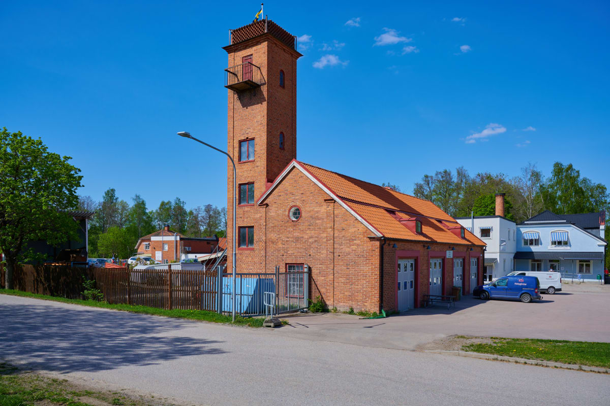 En stor, röd tegelbyggnad med ett högt torn med svensk flagga på toppen, syns under en klarblå himmel. Bredvid finns en lägre byggnadsdel med röda takpannor och garageportar. En asfalterad yta och en gräsbevuxen kant syns framför byggnaden. På höger