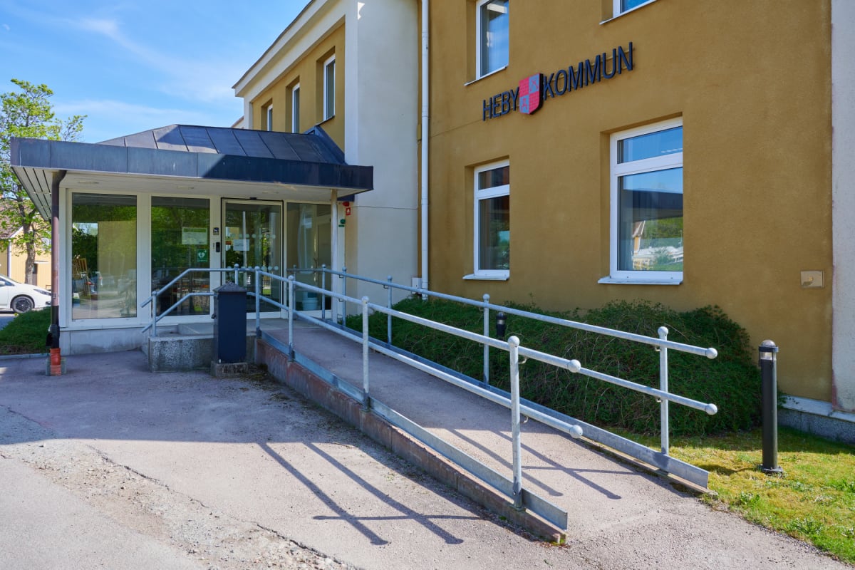 En gul byggnad med skylten "HEBY KOMMUN" ovanför fönster och en entré med en ramp. Gräs och buskar syns runt byggnaden, och en ljusblå himmel med moln ses i bakgrunden. Solen skiner och skapar skuggor på marken.