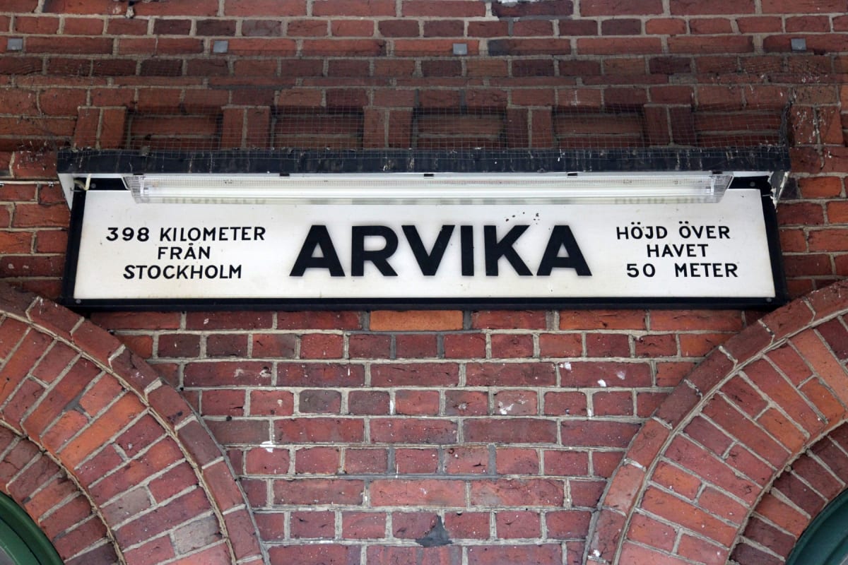 En rektangulär skylt med texten "ARVIKA" i stora svarta bokstäver hänger mot en tegelvägg. Under skylten finns en valvformad tegelkonstruktion. Texten på skylten anger också avståndet till Stockholm och höjd över havet.