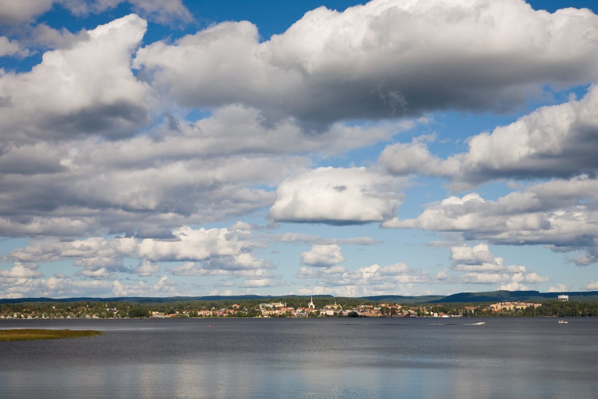 En bild tagen över ett vattenlandskap med en stadsilhuett i horisonten. Himlen är blå med stora vita och grå moln. Vattenytan reflekterar ljuset och på stranden i förgrunden finns vass. En kyrkspirat kan urskiljas i stadsbilden.