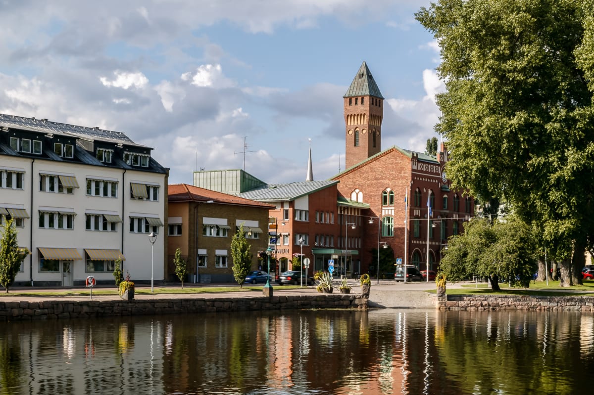 En bild som visar en stadsmiljö med byggnader vid en vattenkant. Byggnaderna varierar i storlek och färg, från vita flerfamiljshus till tegelröda byggnader, inklusive en med ett högt torn. Vattenytan reflekterar byggnaderna och himlen, och längs kant