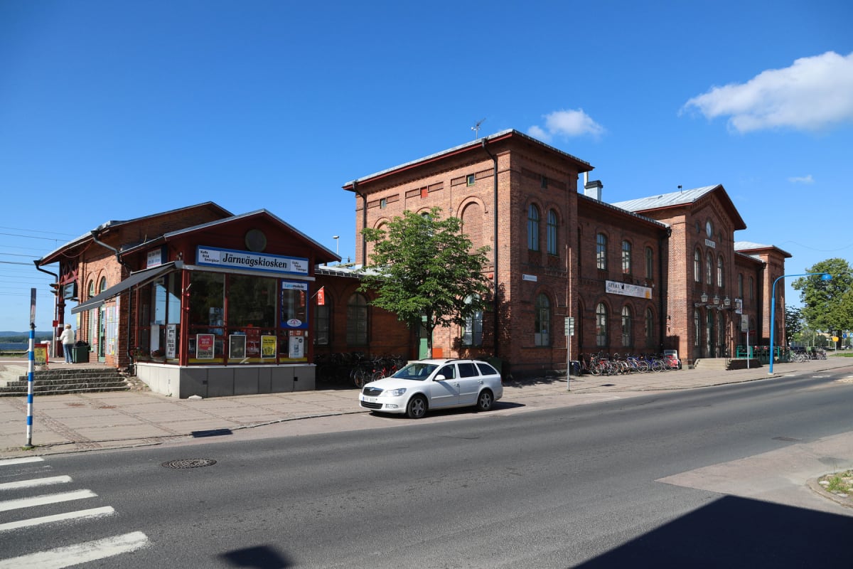 En exteriörbild tagen under dagtid med klart väder, som visar en röd tegelbyggnad vid en gata. Framför byggnaden finns en mindre byggnad med texten "Järnvägskiosken". En vit personbil står parkerad vid trottoaren och cyklar är parkerade intill stora
