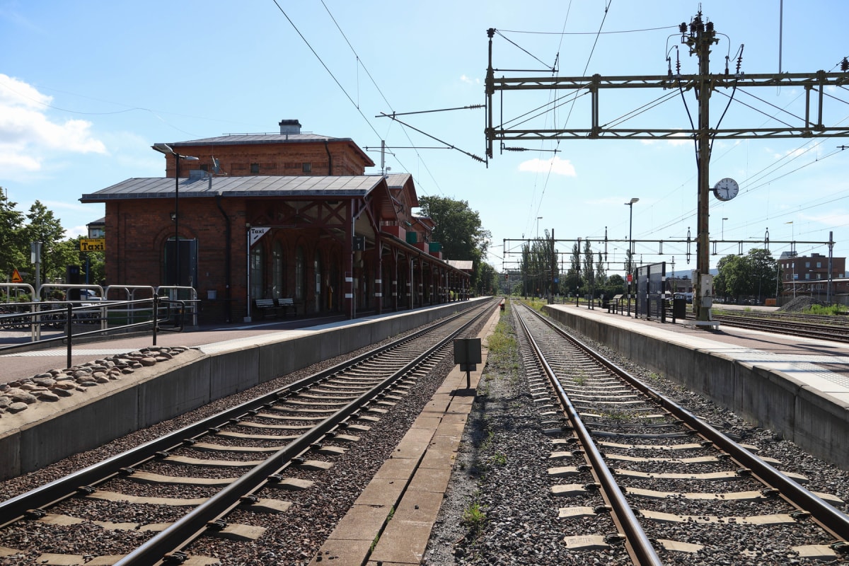 En bild en tågstation med ett stationhus i rött tegel till vänster och två tågspår som sträcker sig in i bilden. En taxiskylt syns tydligt utanför stationhuset. Ovanför spåren finns ett nät av kontaktledningar och till höger skymtar en klocka på en s