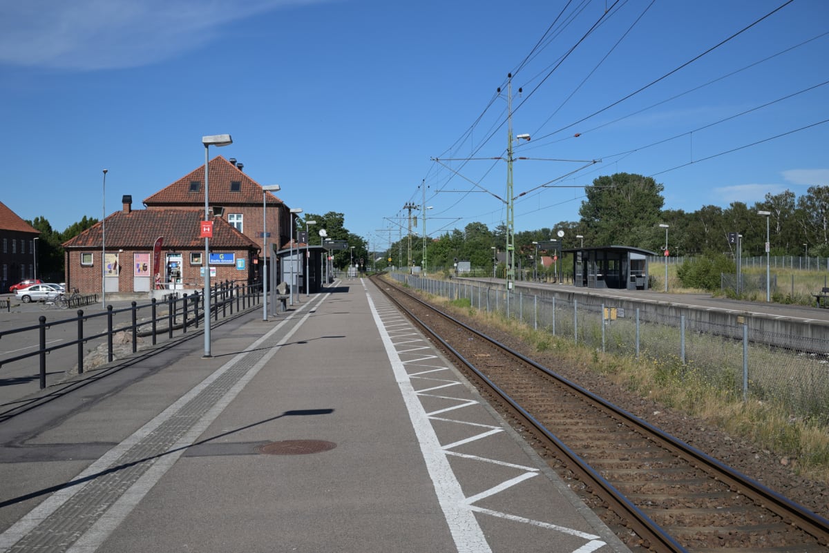En utomhusbild tagen en solig dag som visar en järnvägsstation med ett tegelhus och en perrong till vänster, samt tågspår och en grind i mitten, och ett väderskydd vid en annan perrong till höger. Himlen är klarblå och flera träd ses i bakgrunden.