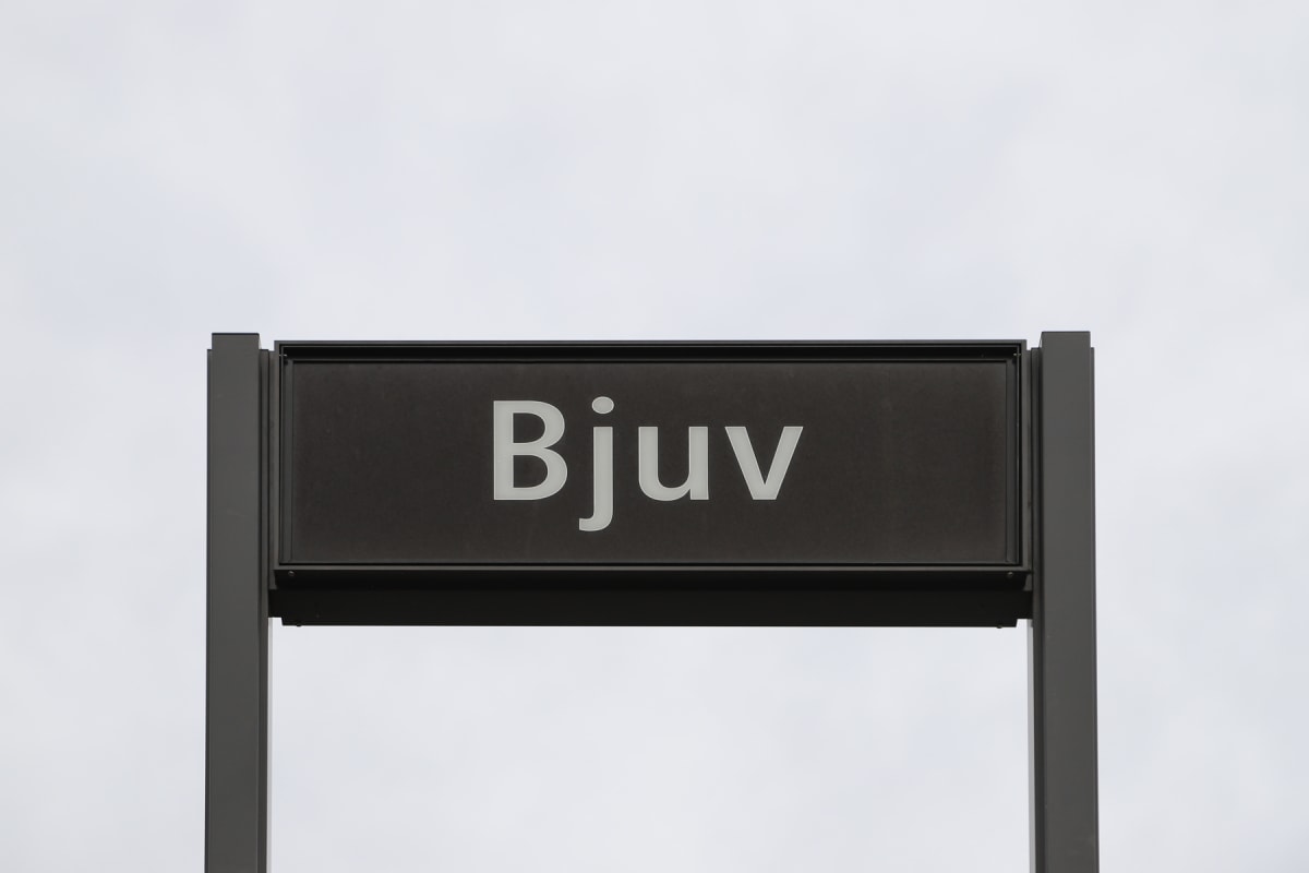 En svart rektangulär skylt med texten "Bjuv" i stora vita bokstäver, monterad på en stolpe, mot en ljusgrå himmel som bakgrund.