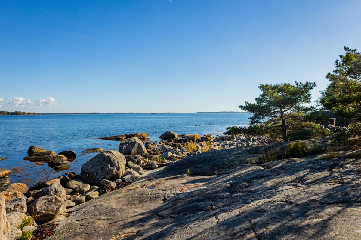 Bilden visar en kustlinje med en stenig strand och lugnt, blått vatten. I förgrunden syns stora klippor och stenar, delvis täckta med alger. Tallar och annan växtlighet växer längs kusten. I bakgrunden syns små öar under en klarblå himmel.