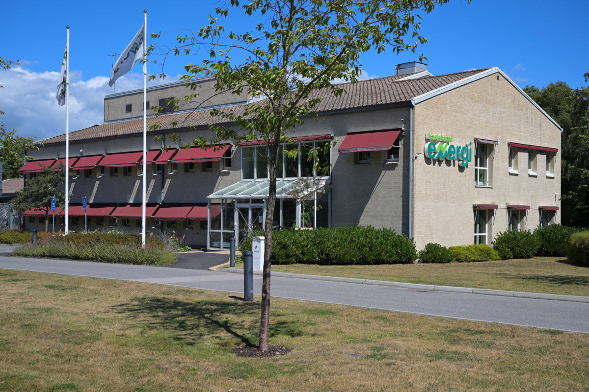 En beige byggnad med röda markiser och ett grönfärgat "Karlshamn Energi"-skylt på väggen. Utanför byggnaden finns en trädgård med gräsmatta, buskar och träd. Två flaggstänger med vita flaggor syns framför byggnaden under en blå himmel med vita moln.