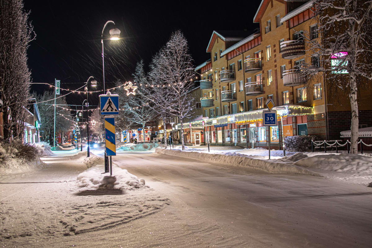 En vinterbild tagen på kvällen eller natten, med snötäckta byggnader och gator. Belysning från skyltfönster, gatlyktor och juldekorationer syns längs en gata med en hastighetsskylt på 30 km/tim och en övergångsställeskylt.
