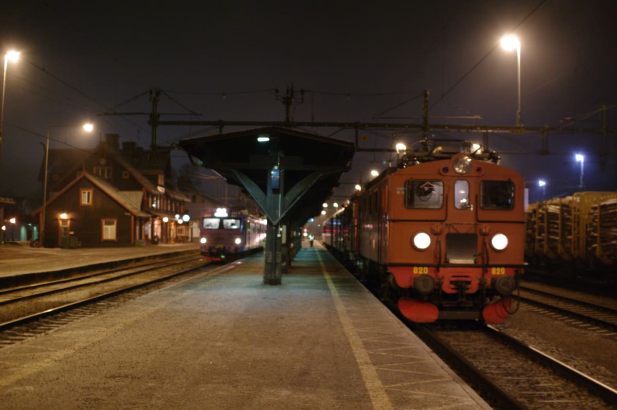En nattbild som visar två tåg på en tågstation. Till höger står ett orangefärgat tåg med nummer 820 synligt. Till vänster syns ett lila tåg. En stationsbyggnad är synlig i bakgrunden. Bilden är tagen utomhus nattetid.