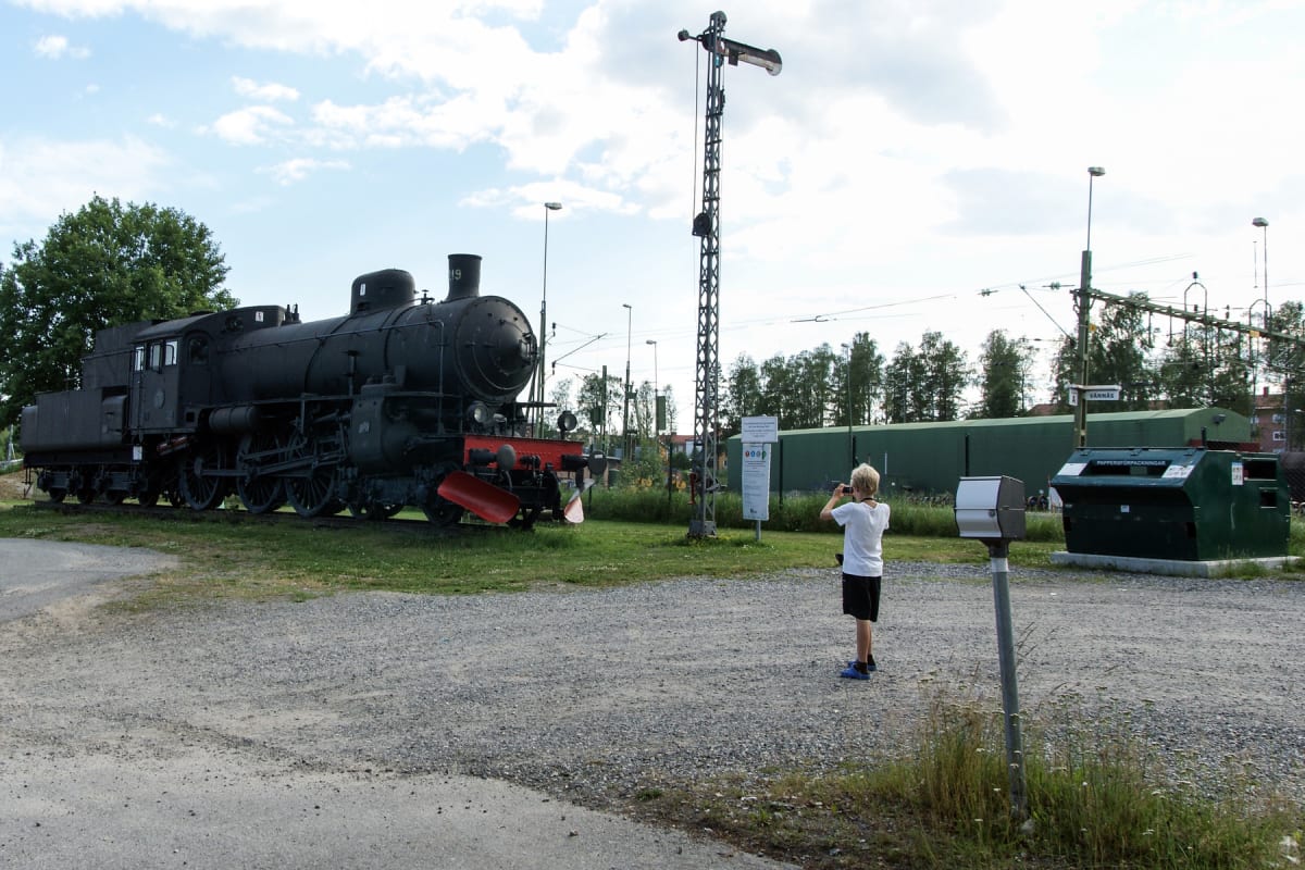 En svart ånglok står på räls bredvid en grusad yta. Ett barn med blont hår fotograferar loket med en mobiltelefon från den grusade ytan. I bakgrunden syns träd, en skylt med texten "PAPPERSFÖRPACKNINGAR" och en grön container. En varningsskylt med te