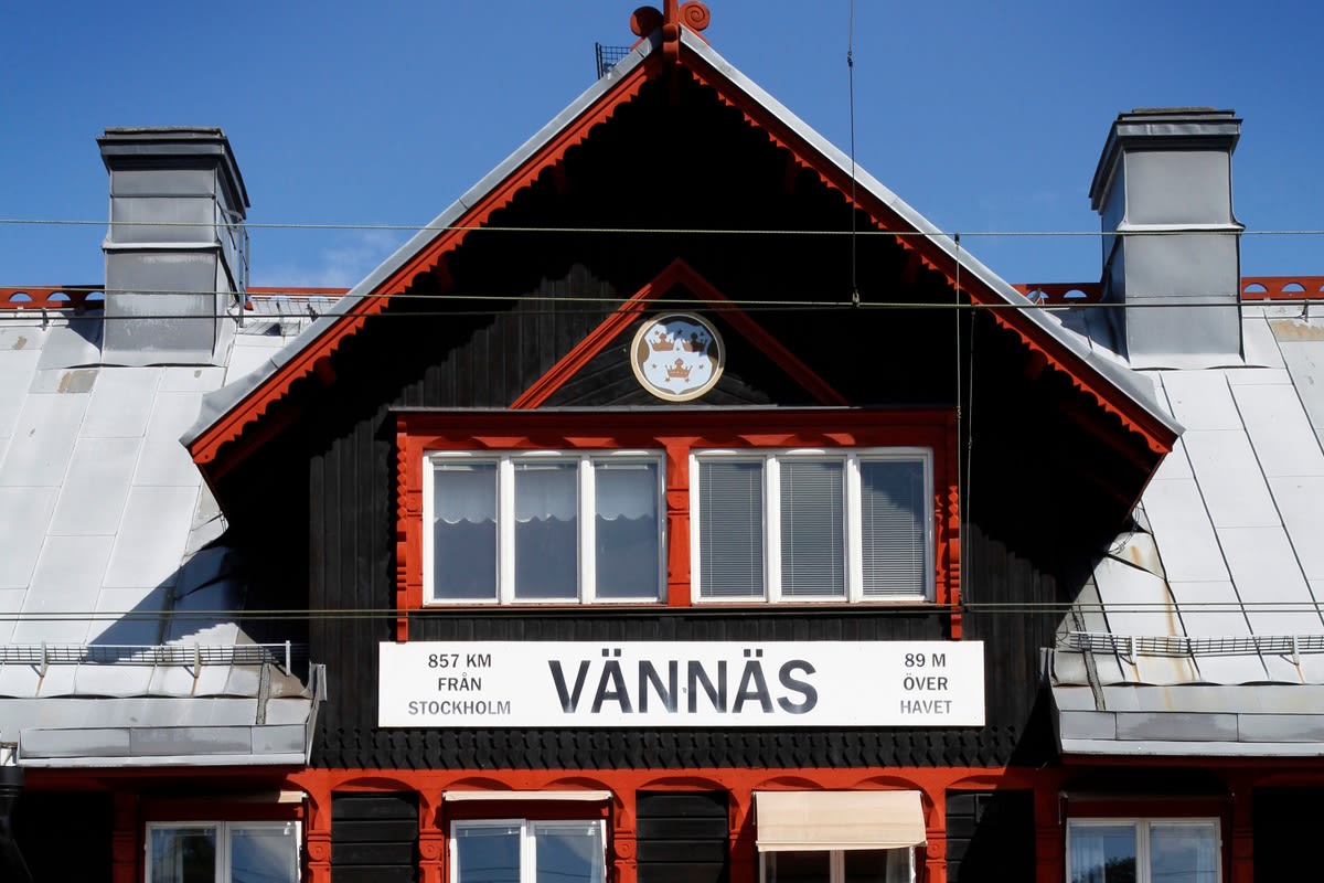 En fasad på en byggnad med svart träpanel, röda detaljer och ett grått tak. En skylt med texten "VÄNNÄS", "857 KM FRÅN STOCKHOLM" och "89 M ÖVER HAVET" syns. Tre fönster med vita karmar och två skorstenar finns på taket.