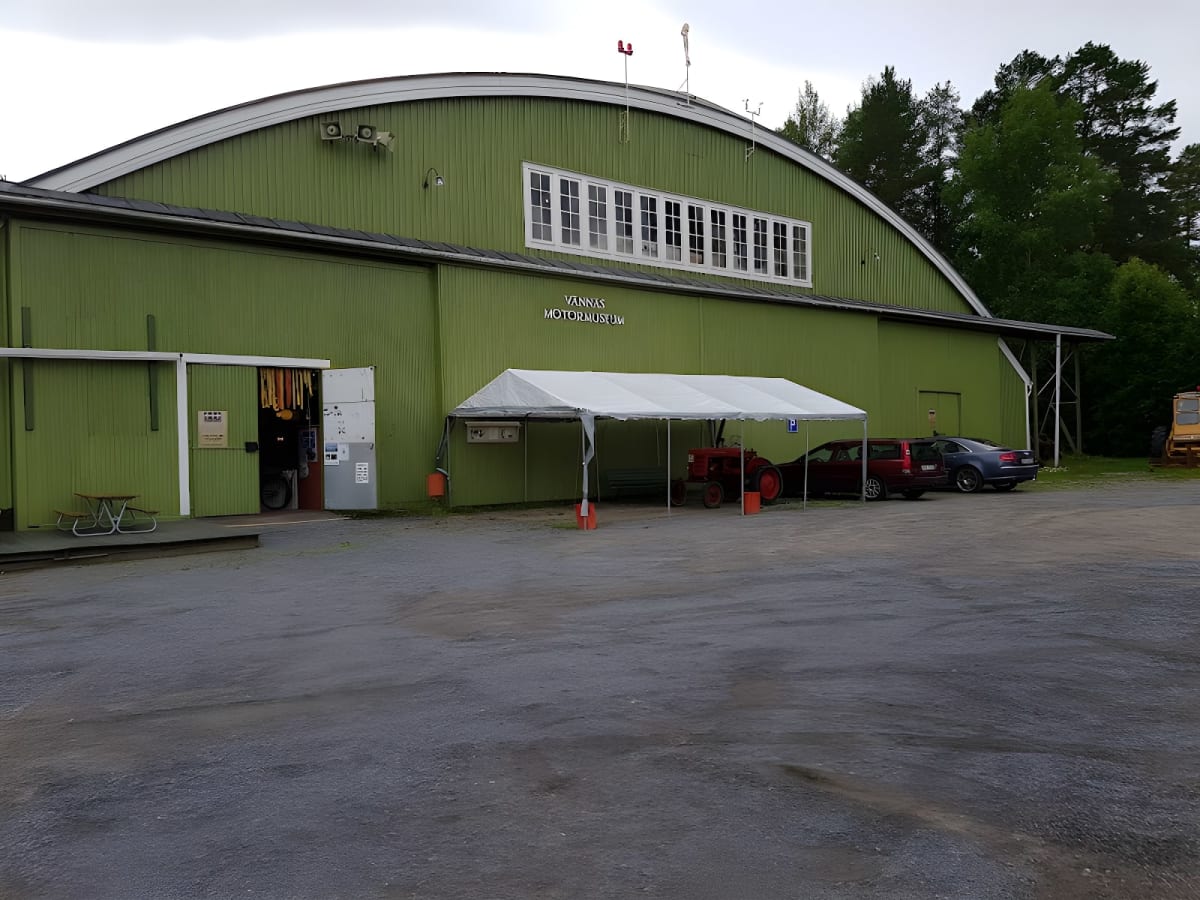 En stor grön byggnad, märkt "Vännäs Motormuseum", står intill en grusplan. Utanför byggnaden finns en vit tältkonstruktion som täcker en röd traktor och en röd bil. En blå bil står parkerad bredvid. I bakgrunden syns träd.