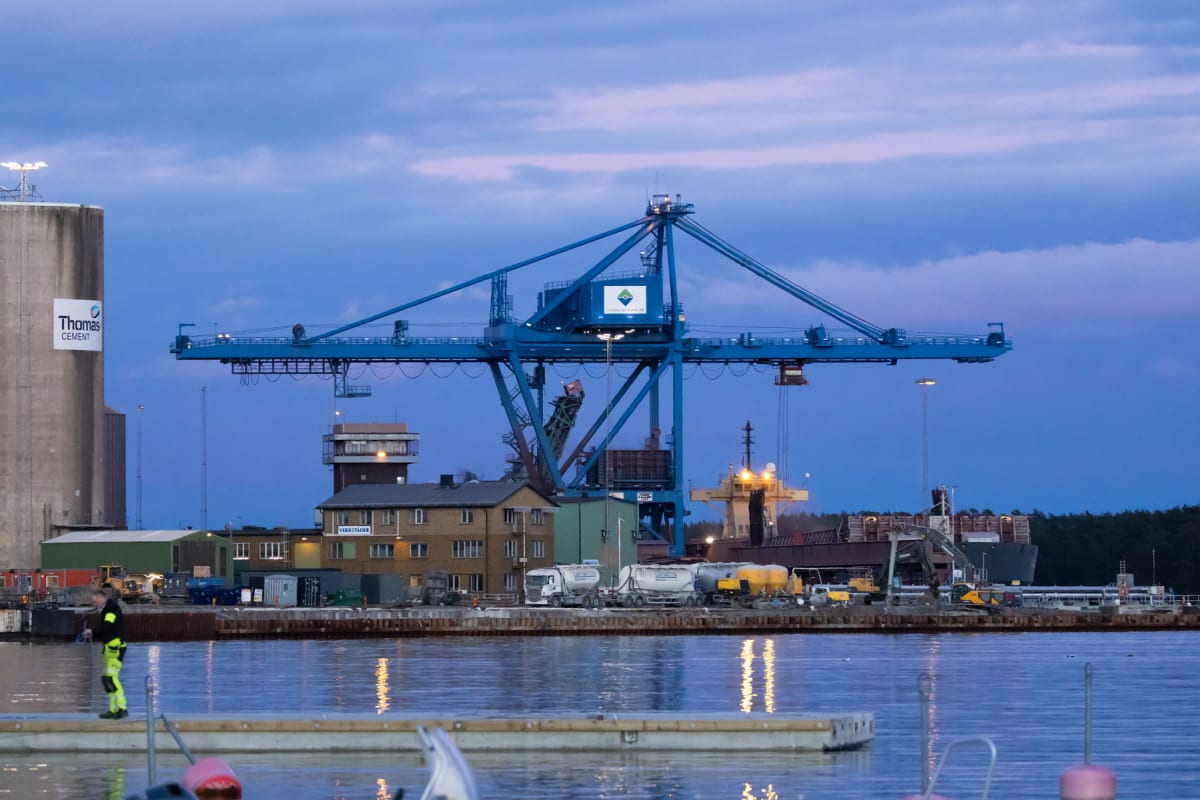 Bilden visar en hamnmiljö i skymningen med en stor blå hamnkran dominerande i mitten. Till vänster syns silos med "Thomas Cement"-logotyp. En person i varselkläder befinner sig på en brygga i förgrunden, och ett vattenområde reflekterar ljus från lan