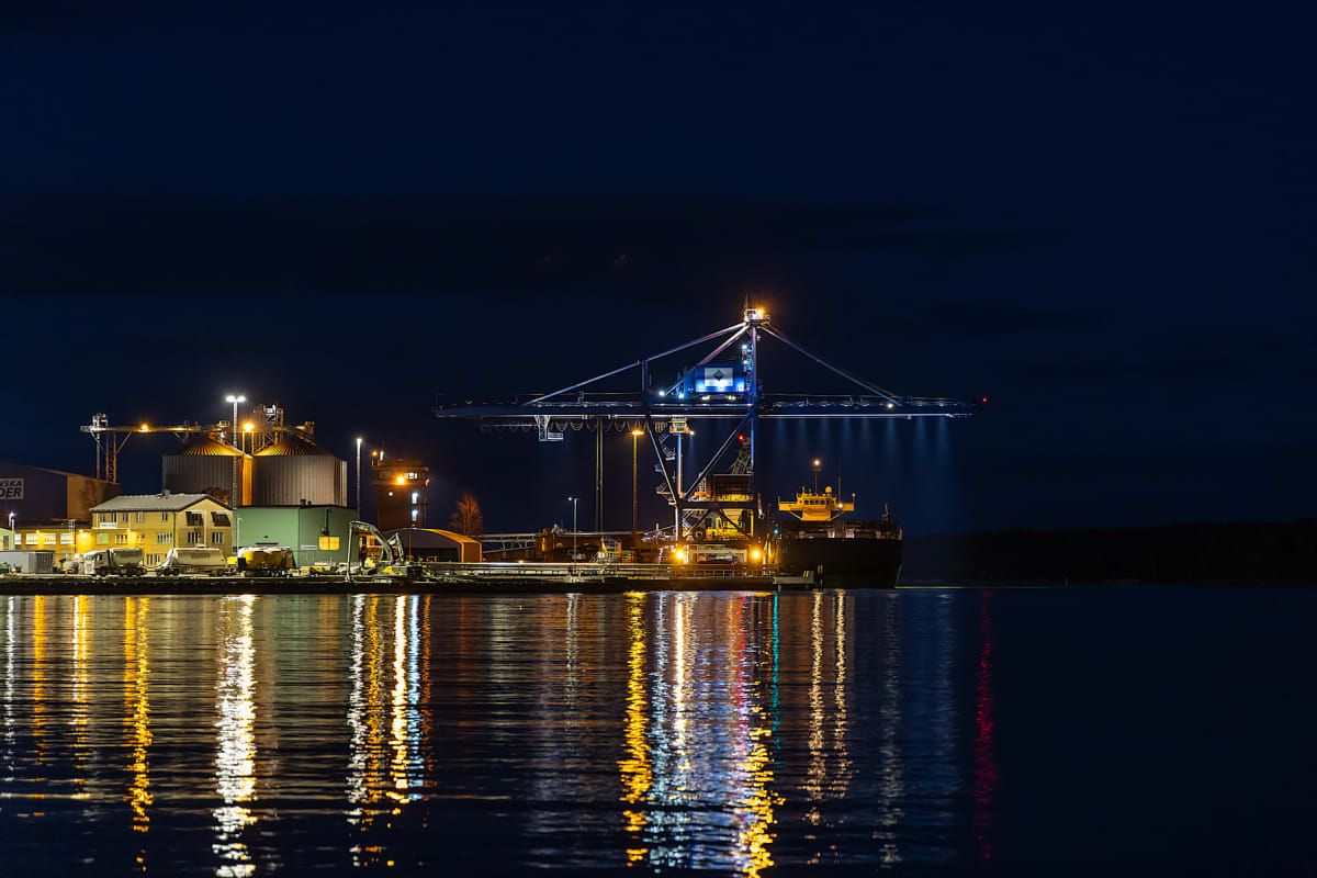 En nattbild över en hamn där silos, byggnader och en kran syns upplysta. Ett fartyg ligger förtöjt vid kajen, och ljusen från land och fartyget reflekteras i vattenytan. Himlen är mörkblå.