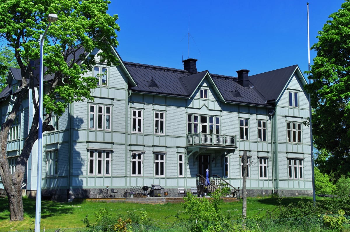 En ljusgrå och ljusgrön träbyggnad med mörkt tak, omgiven av gröna träd och gräs. Byggnaden har många fönster och en balkong på framsidan. Till vänster i bilden syns en gatlykta och en del av ett lövträd.