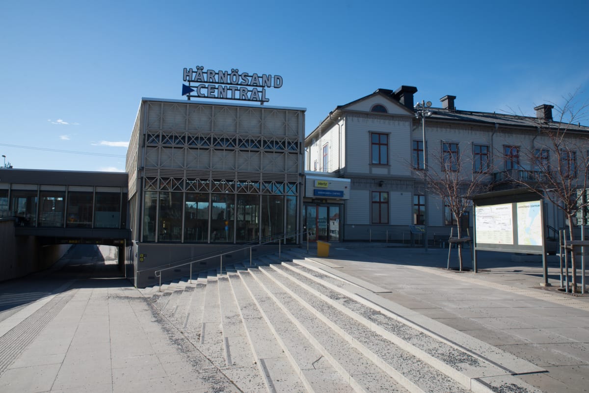 En bild av Häärnösand Central, med en byggnad med skylten "HÄRNÖSAND CENTRAL" överst, en äldre byggnad till höger, en tunnel under en gångbro till vänster, och en trappa i förgrunden. Himlen är klarblå.