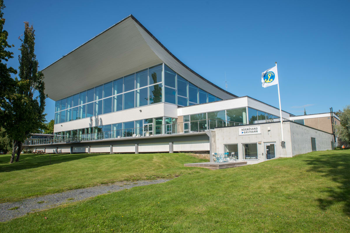 En modern byggnad med stora fönster mot en gräsmatta under en klarblå himmel. På byggnadens sida finns en skylt med texten "HÄRNÖSAND GÄSTHAMN" och en flaggstång med en flagga med logotypen för Härnösand. En grusgång leder till byggnaden, och en träa