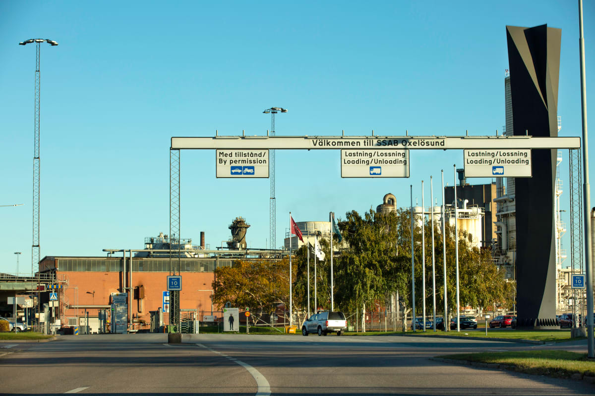 En väg in mot ett industriområde med en stor välkomstskylt för SSAB Oxelösund sträckande sig över vägen. Till vänster syns en tegelbyggnad och master, samt skymtar parkerade bilar. Till höger syns höga industriella strukturer och en svart skulptur.