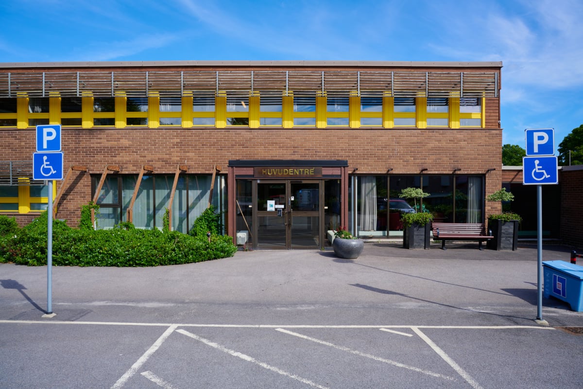 En fasad på en tegelbyggnad med stora fönster, en huvudentré, och två parkeringsskyltar för handikapparkering på en asfalterad yta med parkeringsmarkeringar. Byggnaden har gula detaljer och det är grönska framför den. Blå himmel och solsken.