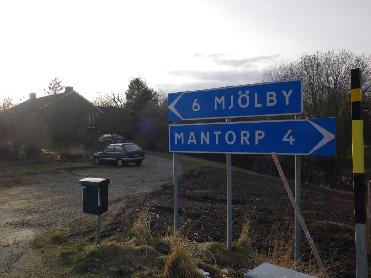 Bilden visar blå vägskyltar som pekar mot Mjölby och Mantorp med avståndsangivelser. I bakgrunden syns ett rött tegelhus, träd utan löv, samt två parkerade bilar på en grusväg. En grön postlåda står i förgrunden till vänster.